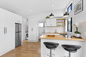 A modern kitchen with white cabinetry, a stainless steel refrigerator, a wooden countertop, and two black bar stools. - Cosy Tiny Home with Ocean Views|by Furnished Rentals (Auckland)