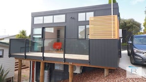 A modern two-story house with a balcony, large windows, and a garage. - Cosy Tiny Home with Ocean Views|by Furnished Rentals (Auckland)