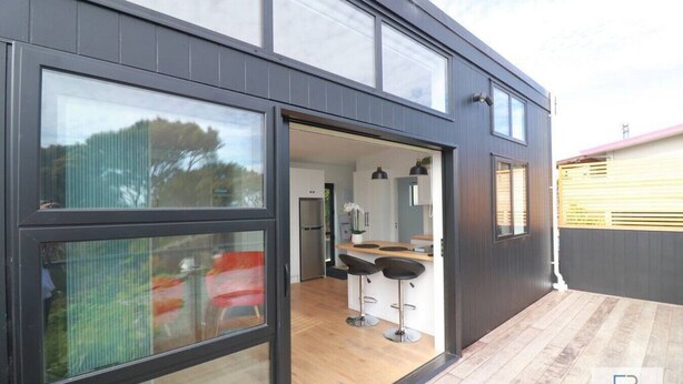 A modern house with a large sliding glass door leading to an open-plan kitchen and dining area. - Cosy Tiny Home with Ocean Views|by Furnished Rentals (Auckland)