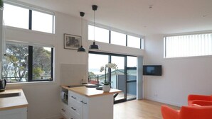 A modern kitchen with a large island, white cabinetry, and a view through the glass door. - Cosy Tiny Home with Ocean Views|by Furnished Rentals (Auckland)