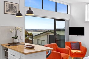 A modern kitchen with a large island, a dining area with orange chairs, and a TV mounted on the wall. - Cosy Tiny Home with Ocean Views|by Furnished Rentals (Auckland)