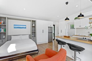 A modern hotel room with a bed, a desk, a kitchenette, and a bathroom. - Cosy Tiny Home with Ocean Views|by Furnished Rentals (Auckland)