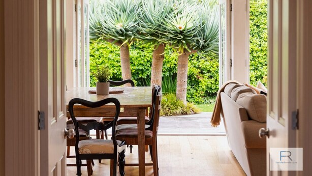 Une porte ouverte donne sur un espace repas comprenant une table et des chaises, un canapé, et une vue sur un jardin avec de hautes plantes et un arbre.