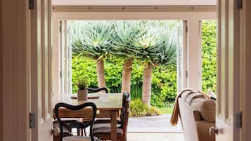 An open doorway leading to a dining area with a table and chairs, a sofa, and a view of a garden with trees and a lawn.
