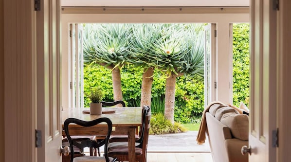A porta aberta dá acesso a uma sala de jantar com mesa e cadeiras, um sofá e vista para um jardim com árvores e gramado.