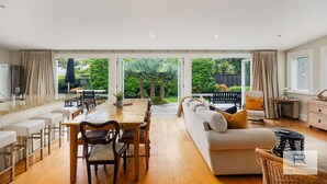 Un vaste espace de vie comprenant une table à manger, un canapé et une vue sur un jardin grâce à de grandes fenêtres.