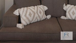 A brown sofa with two patterned throw pillows and a framed picture of a palm tree on the wall.