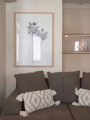 A brown sofa with two patterned throw pillows and a framed picture of a palm tree on the wall.