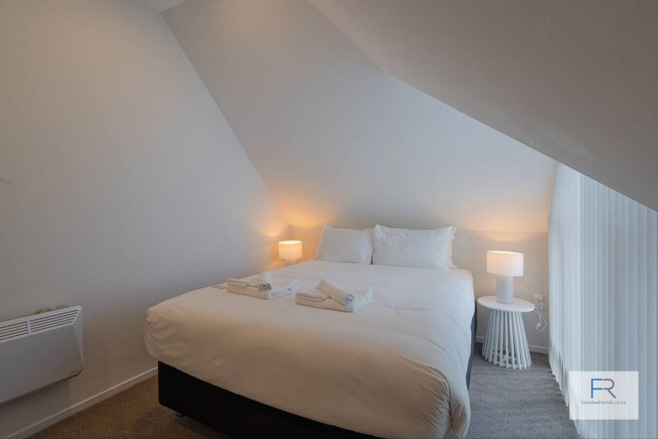 Quarto com uma cama, duas luminárias laterais, um radiador e uma janela com cortinas.