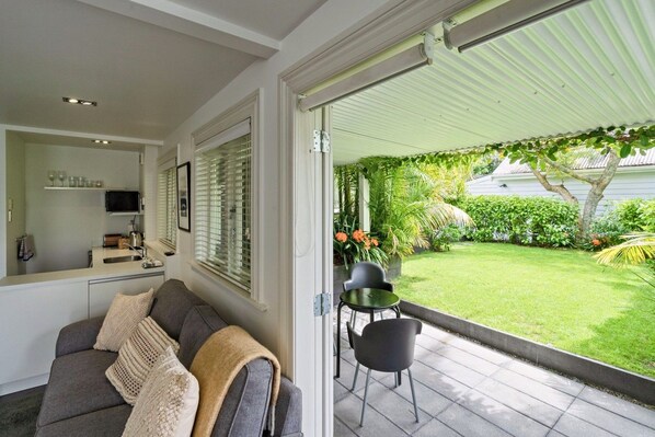 A modern living room with a sofa, a dining table, and chairs, a kitchen area with a sink and a TV, and a view of a well-maintained garden with a lawn and various plants.
