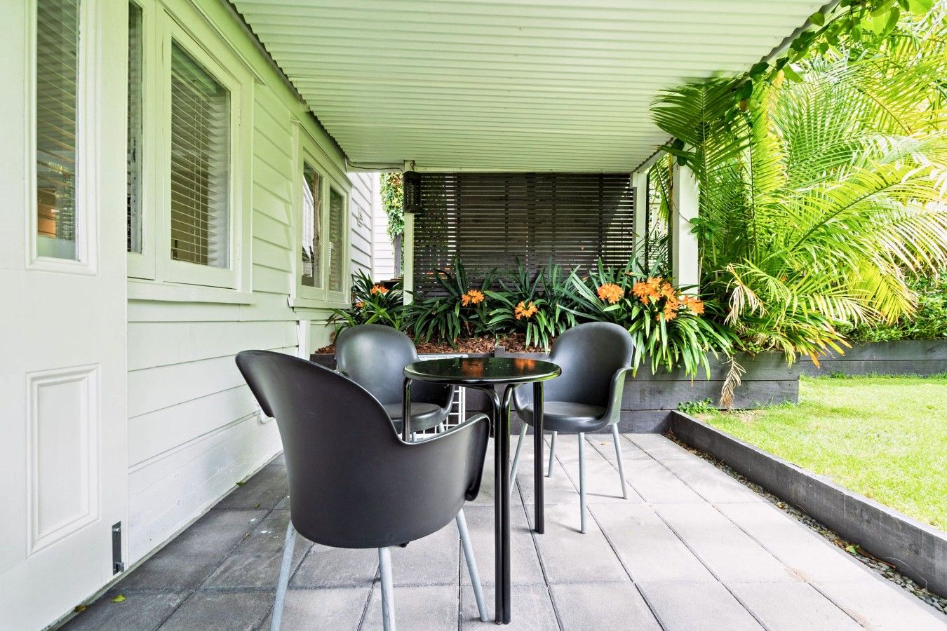 A covered patio with a table and chairs, surrounded by greenery and a window with blinds.