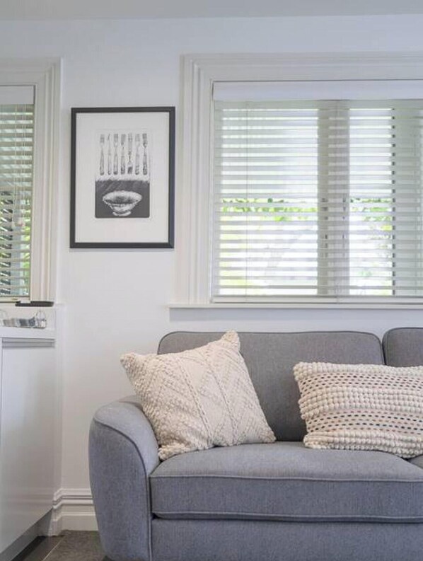 A grey sofa with two textured pillows, a framed picture on the wall, and a window with blinds.