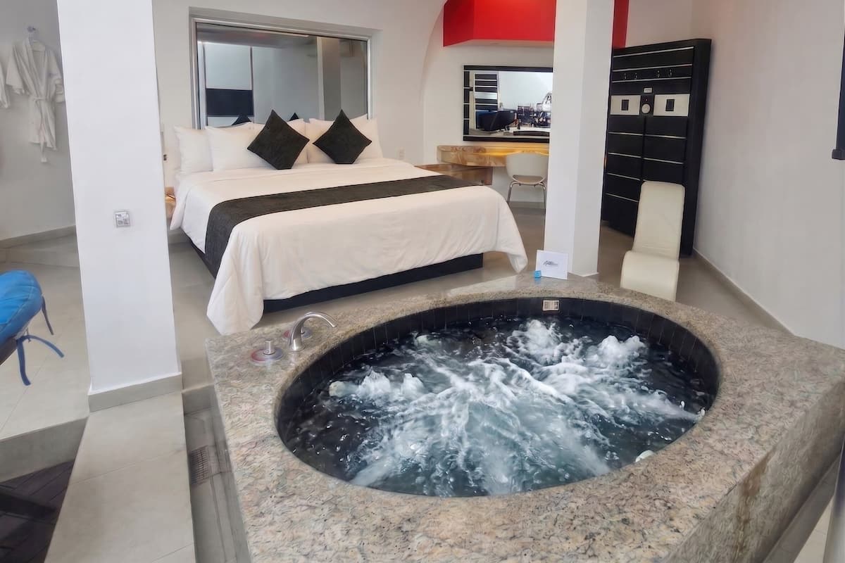 A hotel room with a large bed, a hot tub, a desk, and a view of the city.