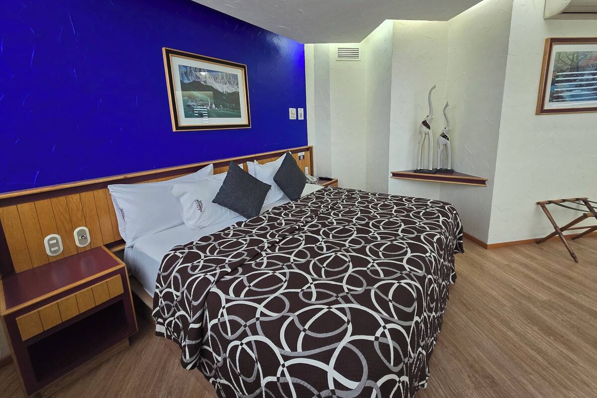 A hotel room with a large bed, a nightstand, a wooden headboard, a framed picture on the wall, and a small shelf with decorative items.