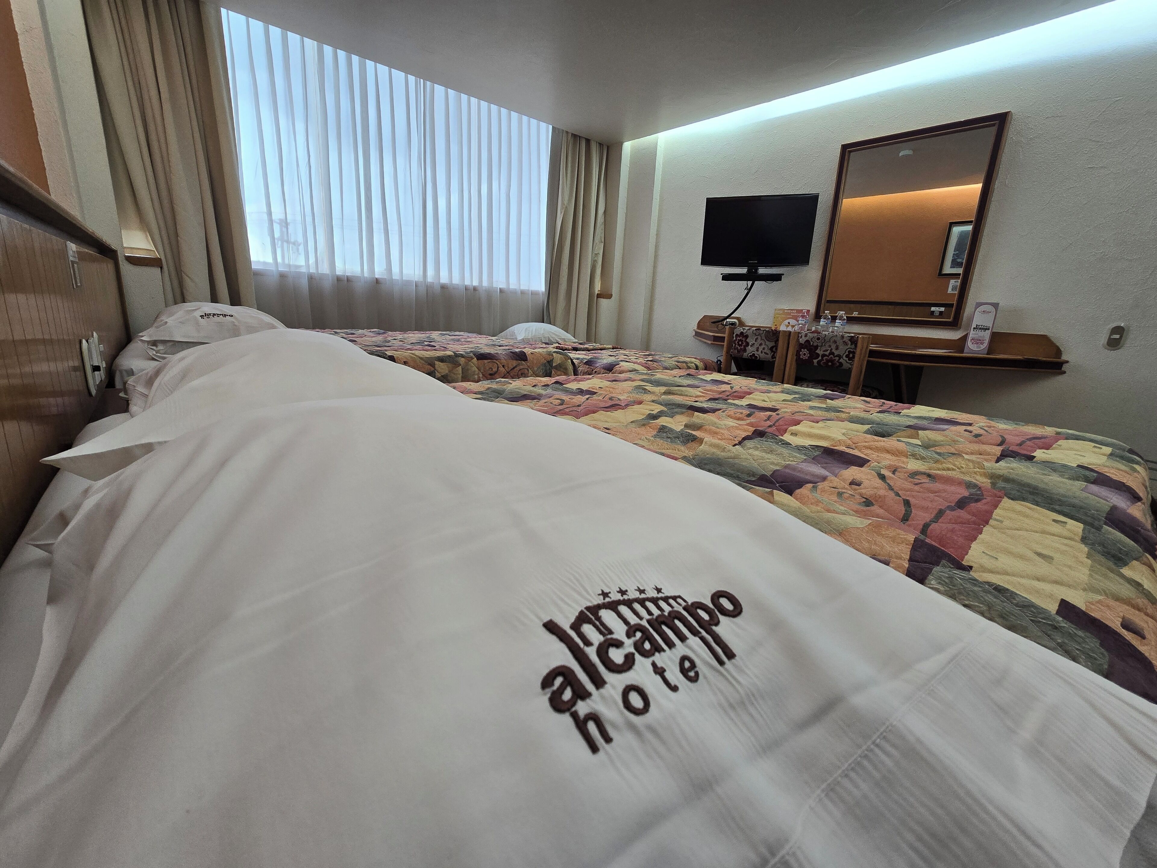 Habitación de hotel con una cama grande, un televisor sobre un mueble bajo, un escritorio con una silla y una ventana con cortinas.