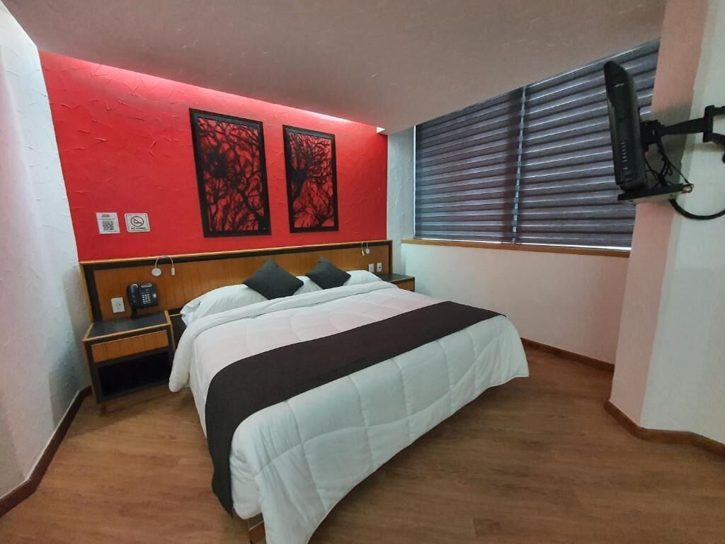 Una habitación de hotel con cama, mesita de noche, teléfono, ventana con persianas y dos obras de arte en la pared.