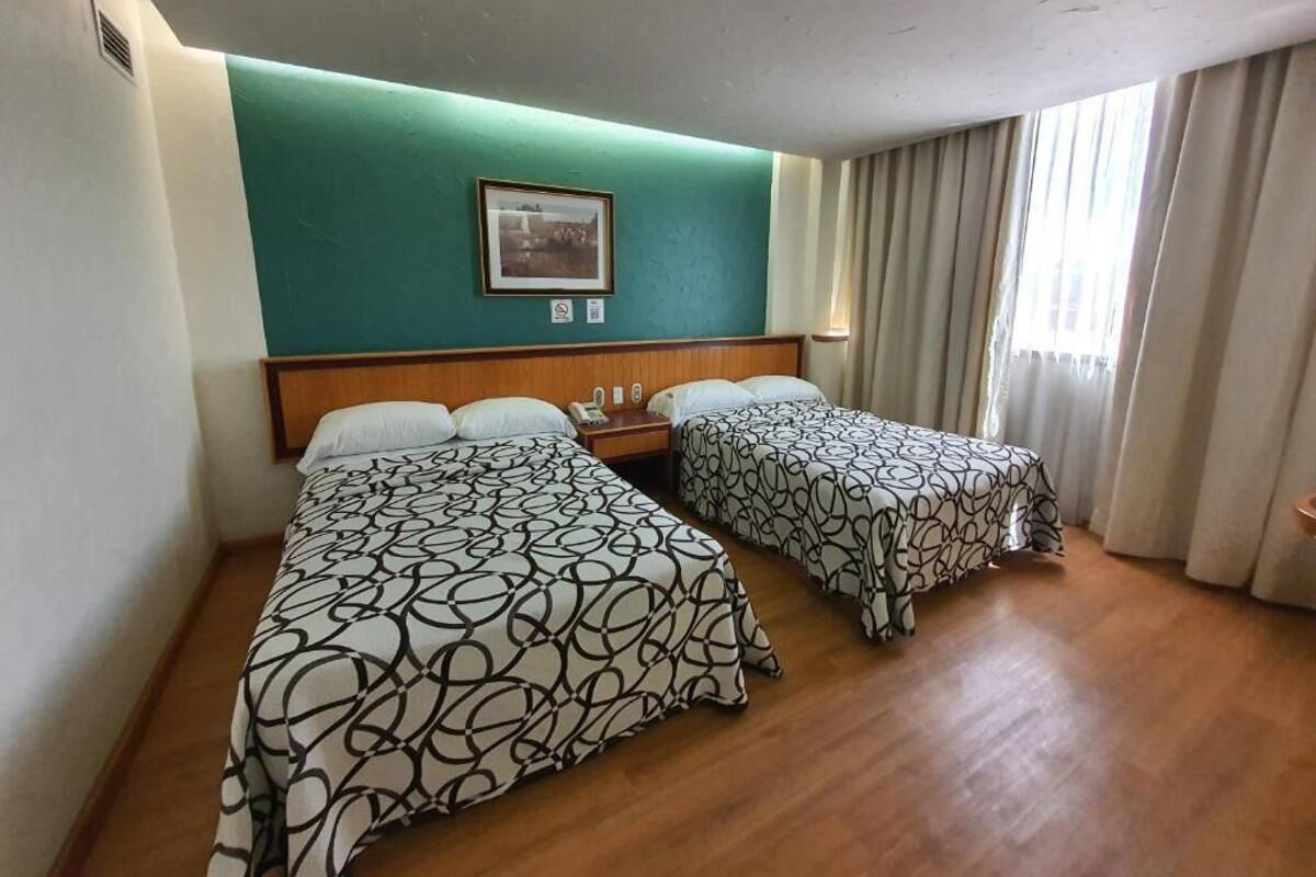 A hotel room with two beds, a teal accent wall, a framed picture, and a window with curtains.