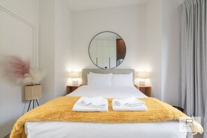 Hotellihuone, jossa on sänky, kaksi yöpöytälamppua, pyöreä peili, yöpöytä lampun kanssa ja ruukkukasvi.
