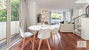 En moderne stue med et rundt spisebord, hvide stole og en sofa. Der er en trappe til højre og et stort vindue til venstre.