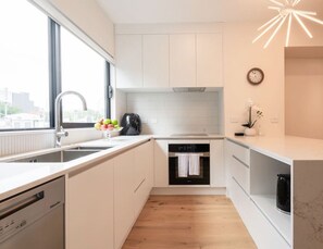 A modern kitchen with white cabinetry, a dishwasher, an oven, and a sink. There is a window providing natural light and a decorative light fixture above the counter.