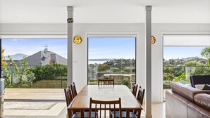 A modern dining area with a large table and chairs, large windows offering a view of a residential neighborhood and hills. - Stunning Poolside Residence| by Furnished Rentals (Auckland)