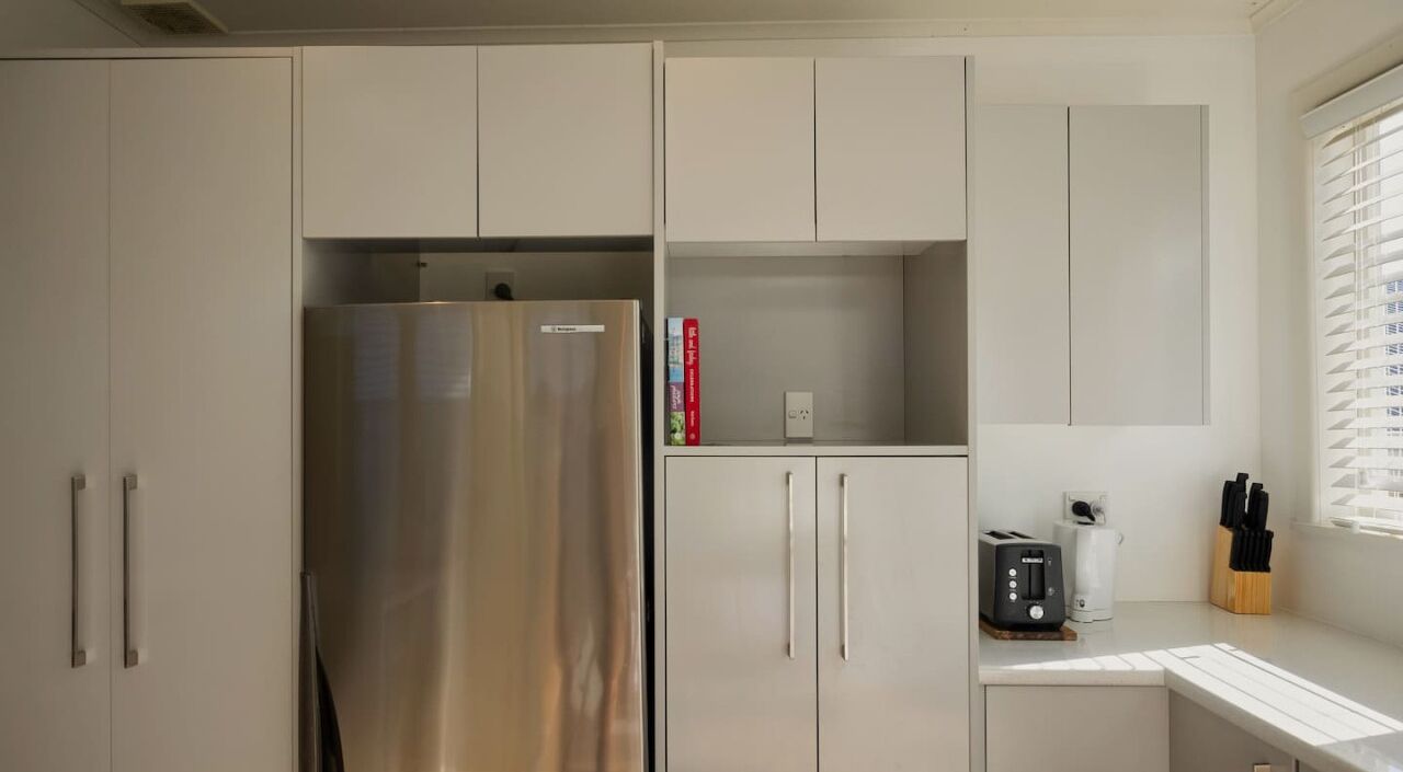 A modern kitchen with a stainless steel refrigerator, white cabinets, and a window with blinds.
