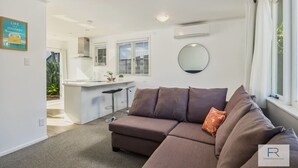 Ein modernes Wohnzimmer mit einem braunen Ecksofa, einer Küchenzeile mit einem Barhocker und einem großen Fenster mit Vorhängen.