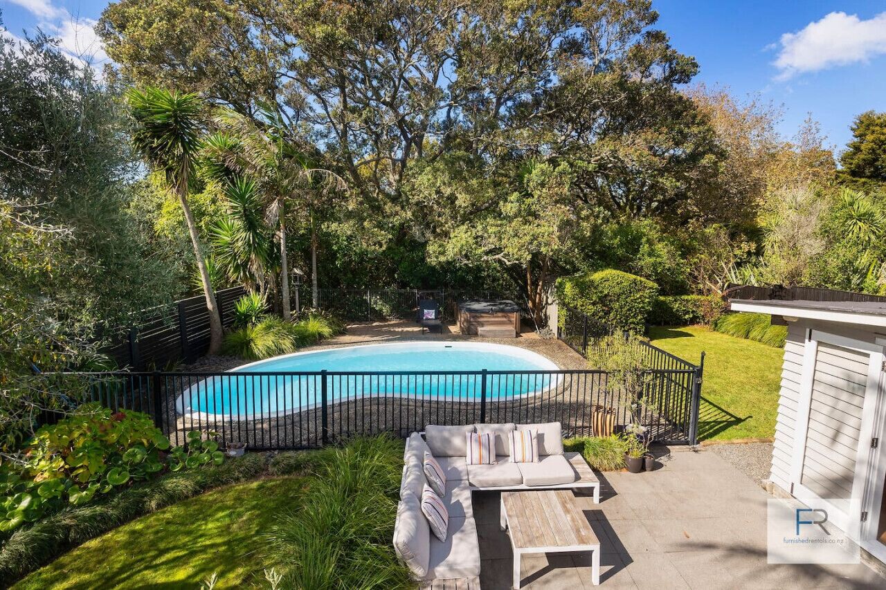 A backyard with a swimming pool, surrounded by a black fence, and featuring a wooden deck with a sofa and a small shed.