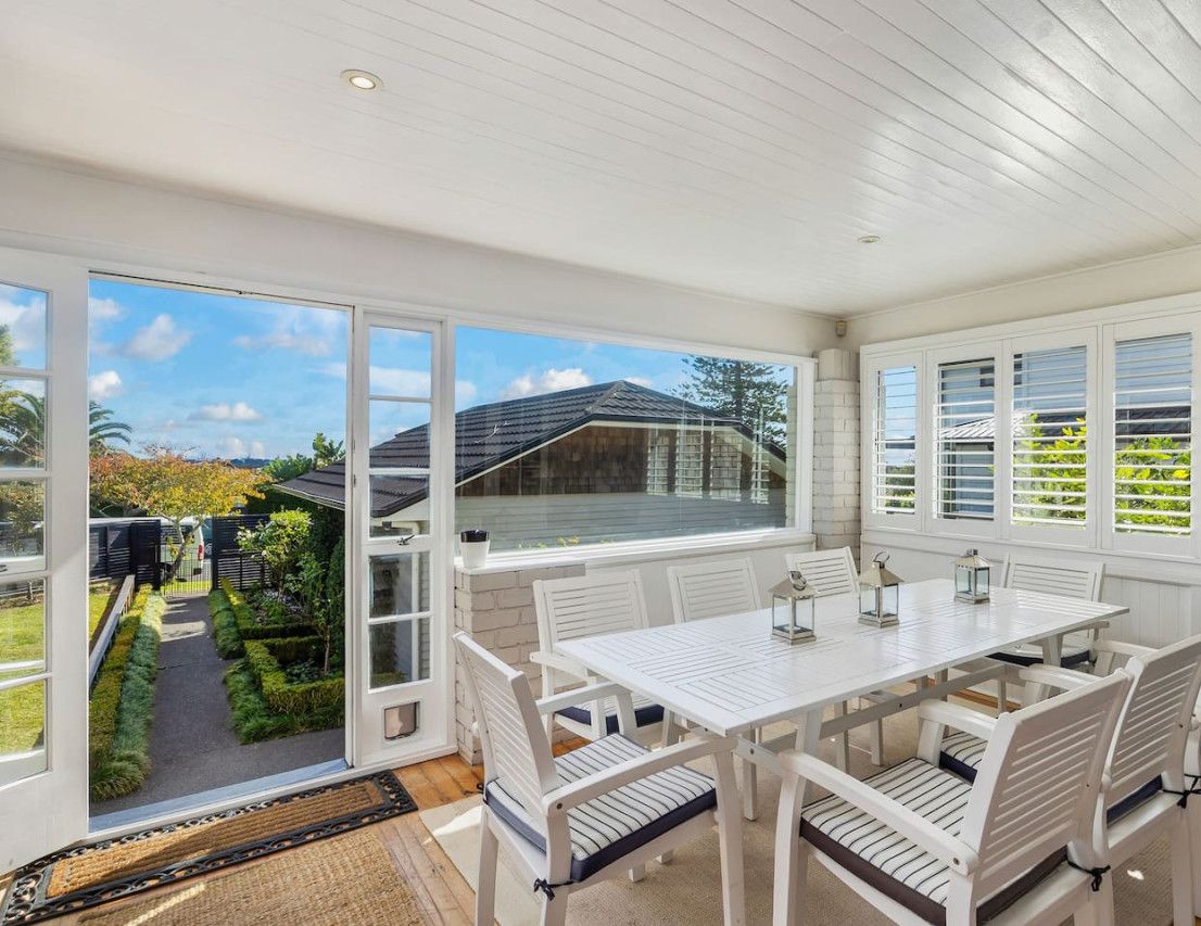 A dining area with a white table and chairs, large windows, and a view of a garden and neighboring houses.