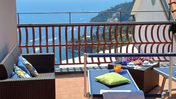 Un balcon agrémenté de meubles en osier, une table avec des boissons et des assiettes, et une vue sur la mer et les collines.