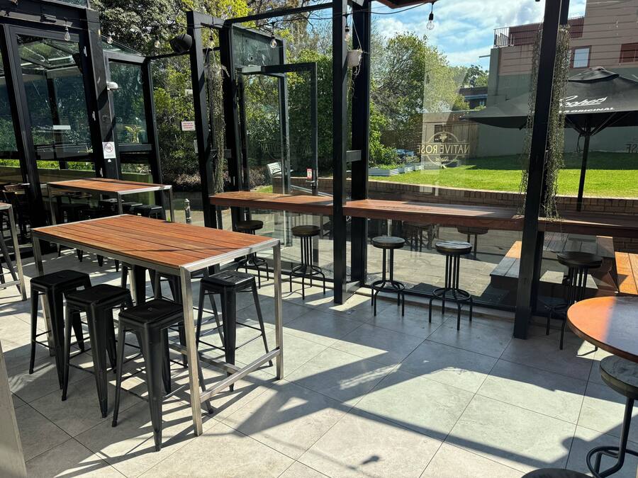 An outdoor seating area with tables and stools, surrounded by glass walls and metal frames.