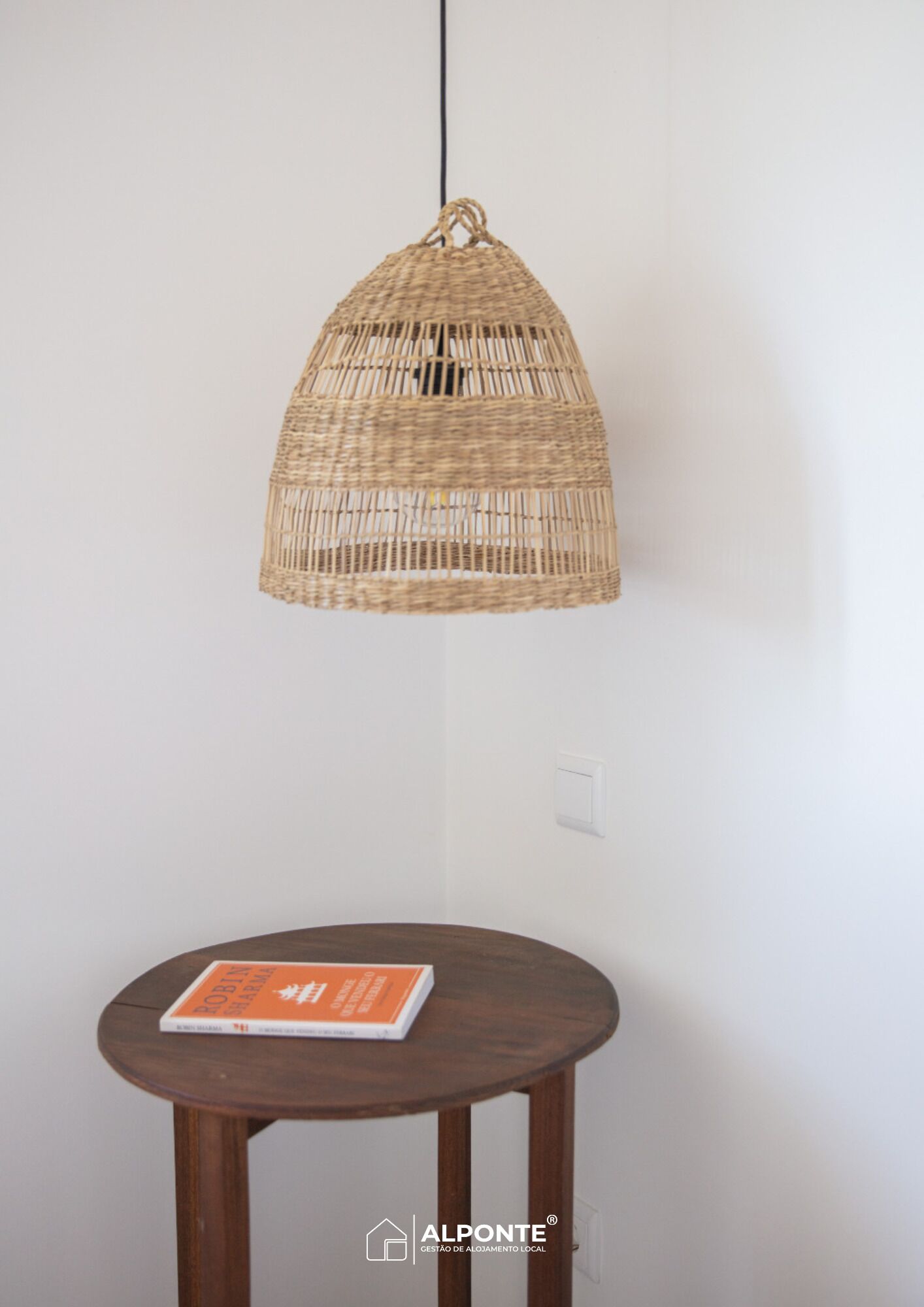 Una lámpara colgante de mimbre suspendida del techo, una mesa redonda de madera con un libro, y una pared blanca con un interruptor de luz.