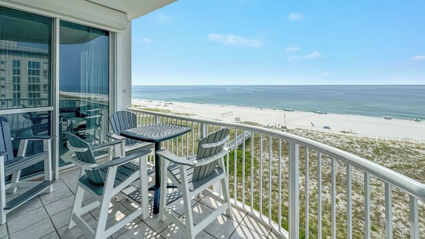 A balcony with a table and chairs, overlooking a beach and ocean.