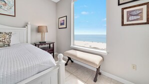 A bedroom with a bed, a bench, a nightstand, a lamp, and a window with a beach view.
