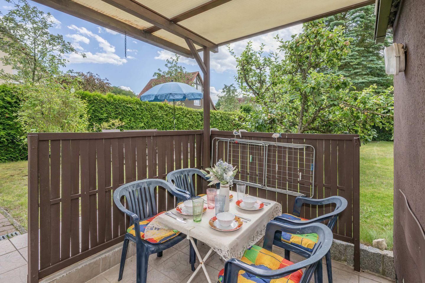 Eine Terrasse im Freien mit einem Tisch für zwei Personen, umgeben von Stühlen mit bunten Kissen, einer weißen Tischdecke und einer kleinen Vase mit Blumen.