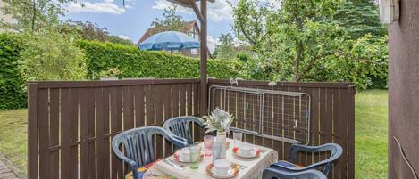 Eine Terrasse im Freien mit einem Tisch für zwei Personen, umgeben von Stühlen mit bunten Kissen, einer weißen Tischdecke und einer kleinen Vase mit Blumen.