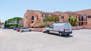 Un estacionamiento con una camioneta blanca, un coche rojo y un coche plateado estacionados frente a un edificio con balcones y un cantero de flores rosa.