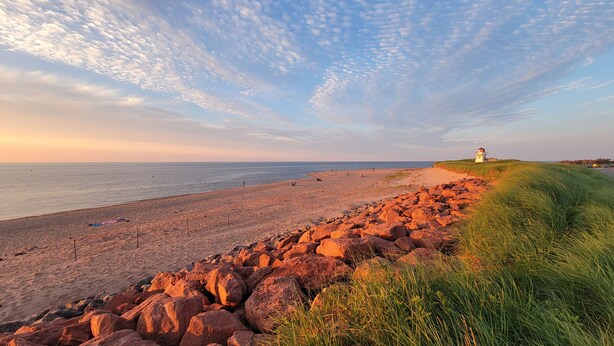 En strand med en stenig kustlinje, en grässtig och en fyr i fjärran.