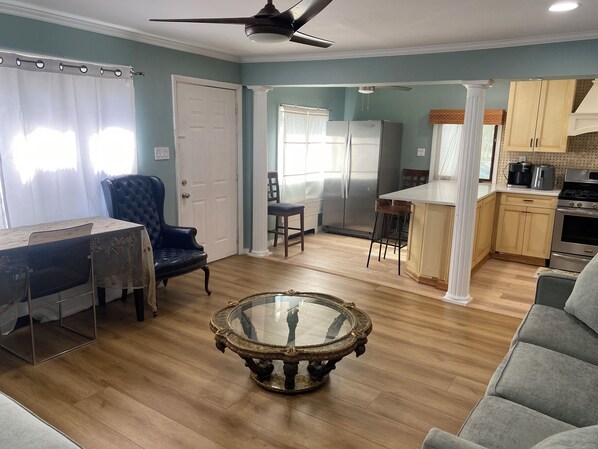 A living room with a glass coffee table, a dining area with a chair, a kitchen with cabinets and appliances, and a ceiling fan. - 2 Bedroom Apartment Near UBS Arena, JFK, & NYC (Elmont)