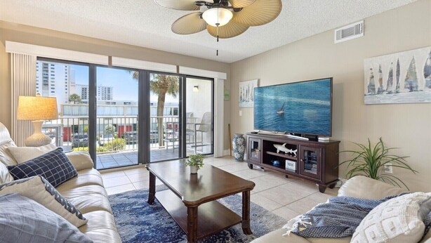 A spacious living room with a large TV, comfortable seating, a ceiling fan, and a balcony with a city view.