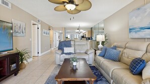A spacious living room with a beige sofa, a coffee table, and a ceiling fan. There is a television on a stand and a plant on the floor.