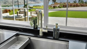 A kitchen with a stainless steel sink, a faucet, and a view of a landscaped garden through a window. - Luxury 4 BR w/ Serene Lake Views (Spring Valley)