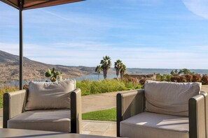 An outdoor seating area with two cushioned chairs and a table, overlooking a scenic landscape with hills and a body of water.