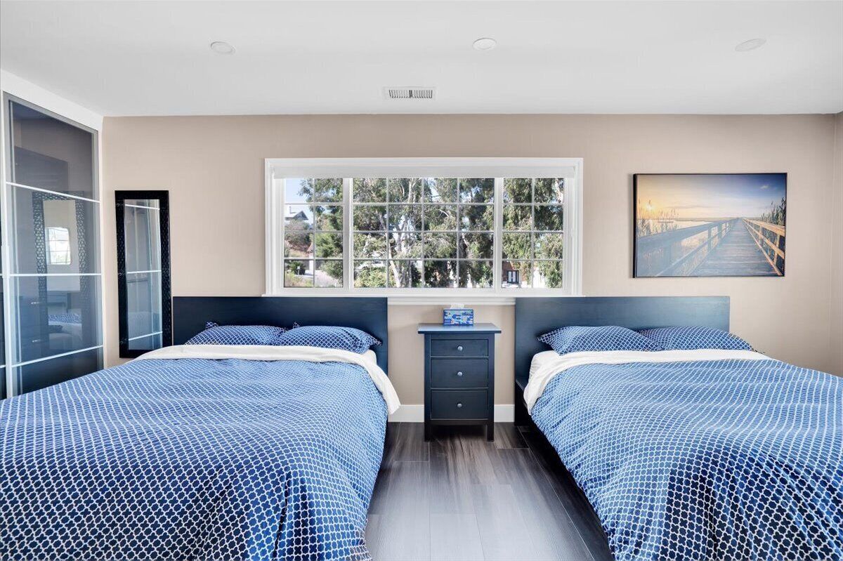 Un dormitorio con dos camas, una ventana que da a árboles, una mesita de noche y un cuadro en la pared.