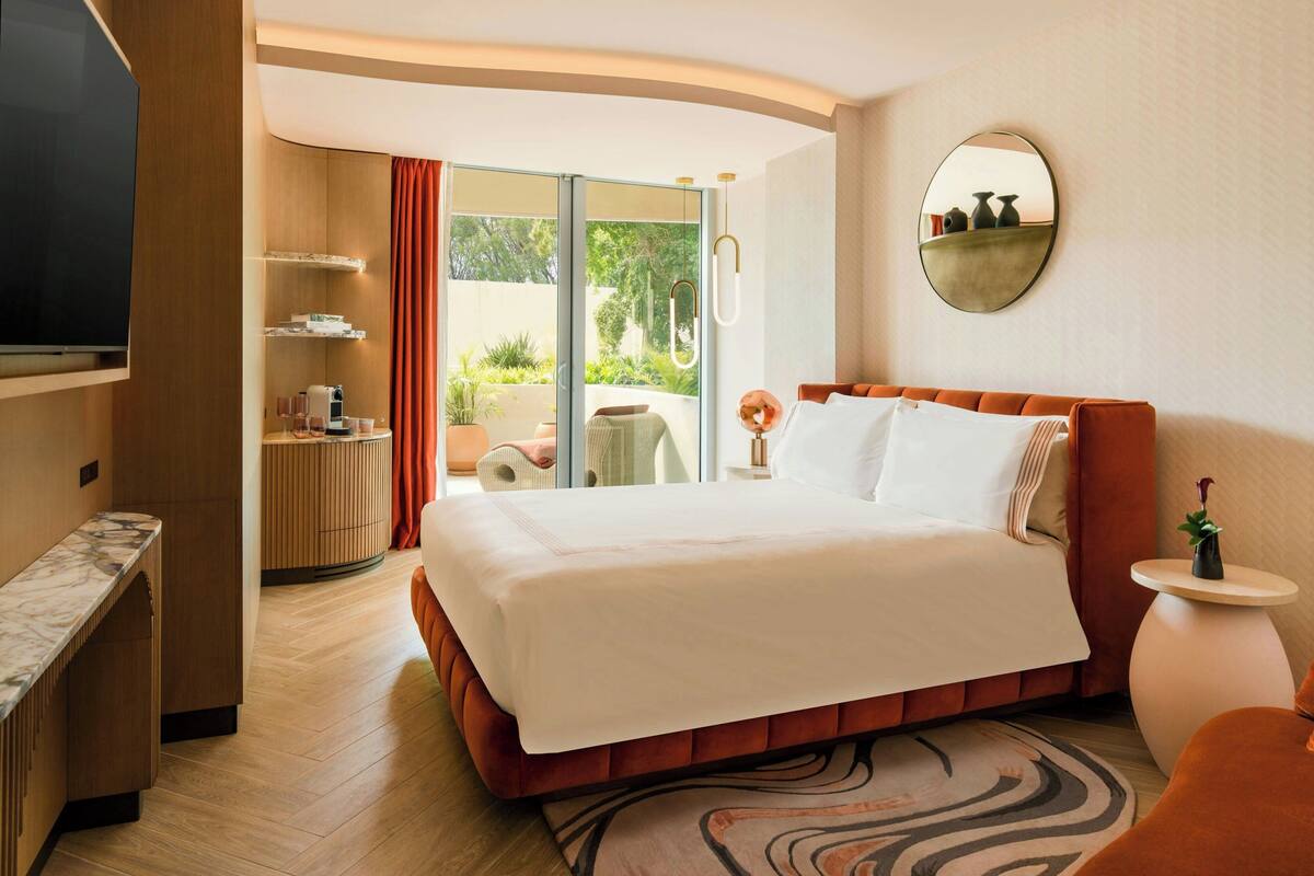 A hotel room with a bed, a TV, a desk, a round mirror, and a view of the outdoors.