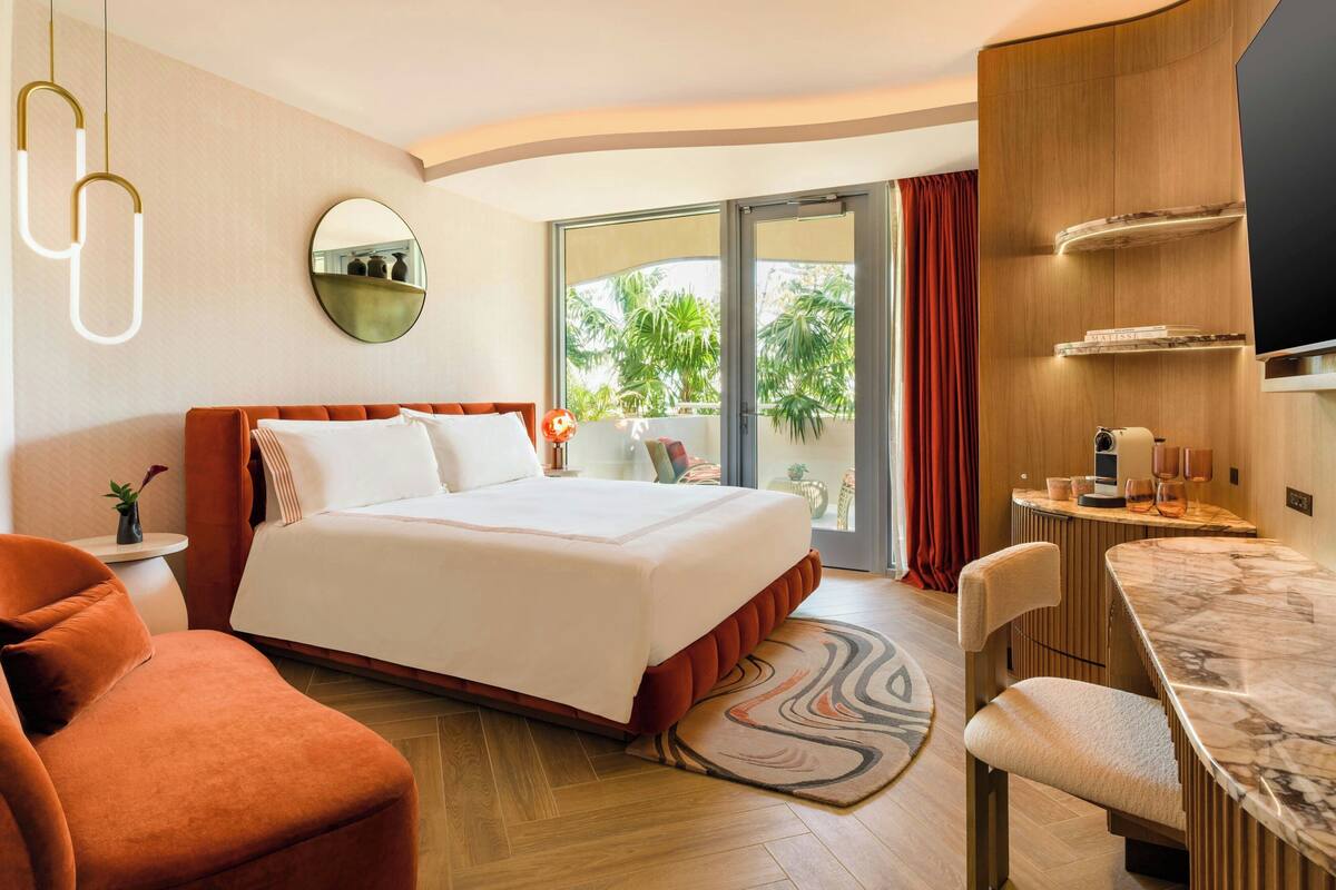 A modern hotel room with a bed, a seating area, a TV, and a balcony with a view of palm trees.