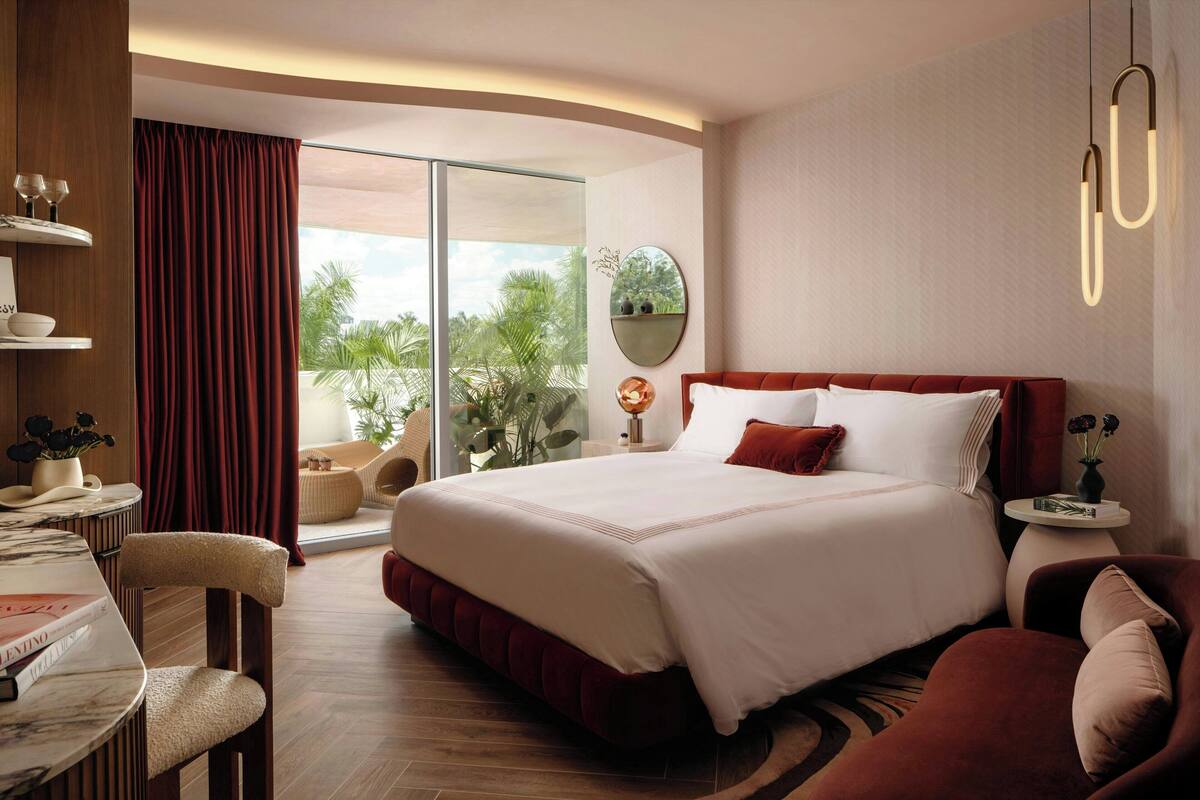 A hotel room with a large bed, a desk with a chair, a round mirror, and a view of the outdoors.