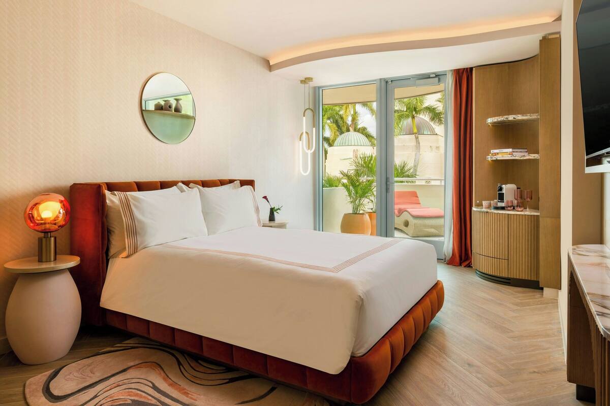 A hotel room with a bed, a bedside lamp, a mirror, a TV, a small table with a plant, and a view of the outdoors.