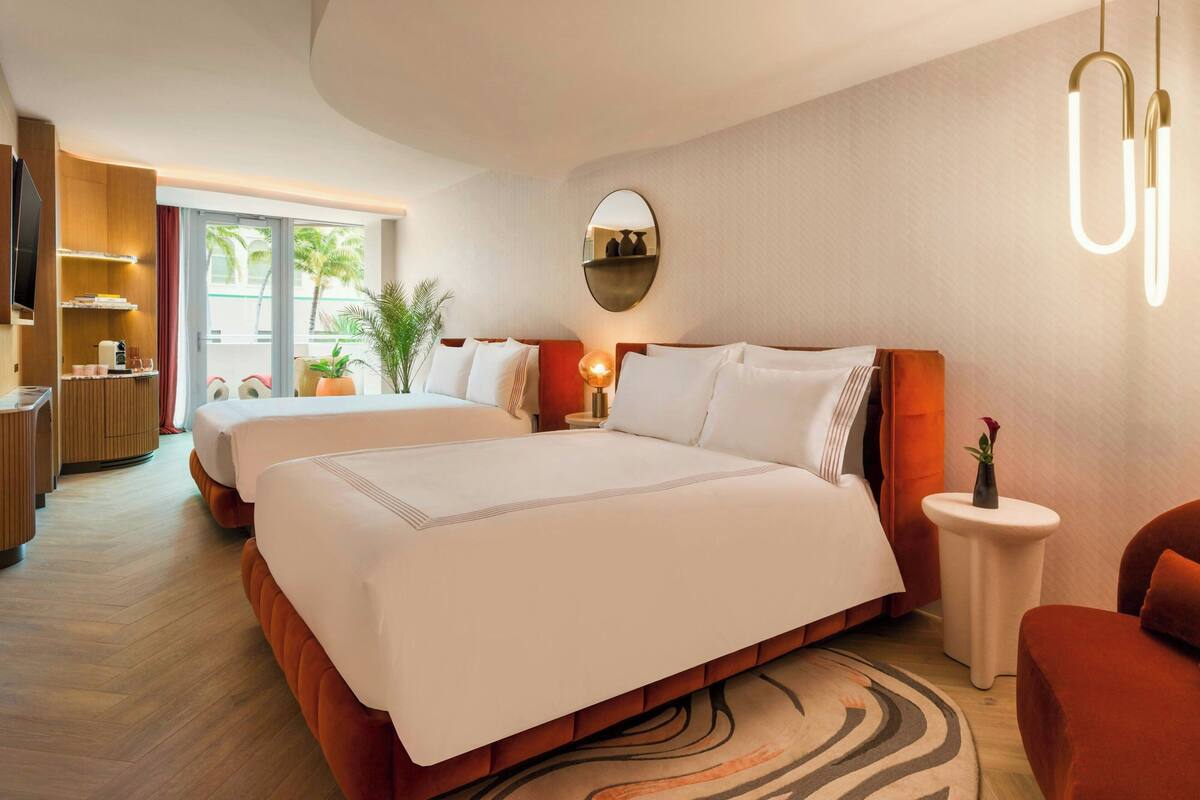 A hotel room with two beds, a TV, a desk, and a view of palm trees.