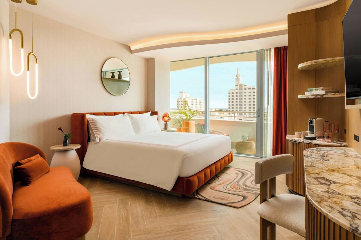 A hotel room with a bed, a chair, a table, a TV, and a view of buildings.